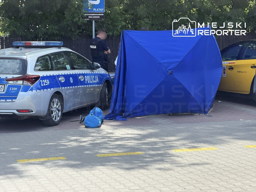 Policjant rozmawia z kolegą obok radiowozu, a w tle znajduje się niebieski namiot oraz torba.