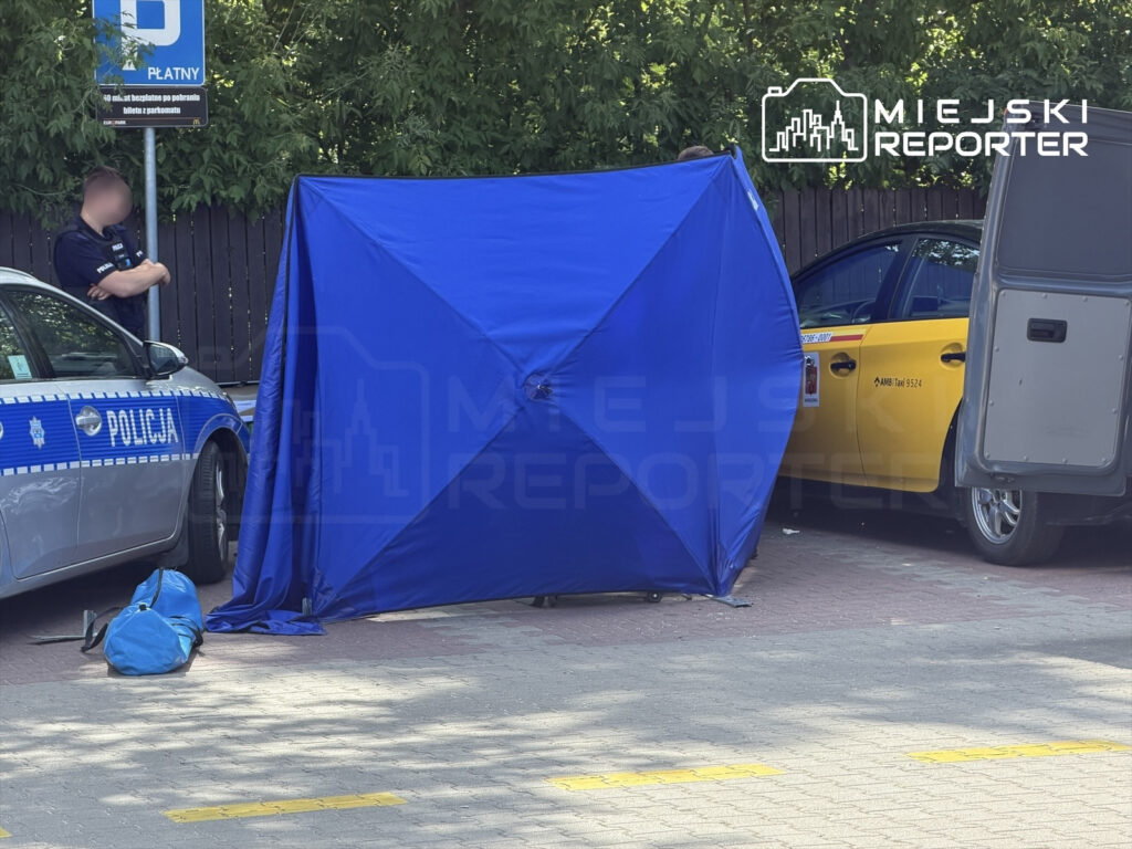Policjant stojący obok nieoznakowanego policyjnego samochodu, obok niebieskiego namiotu na parkingu.
