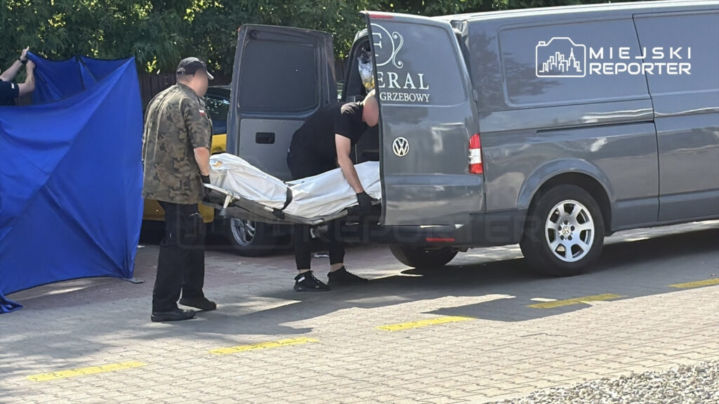 Mężczyźni w mundurach przenoszą ciało na noszach do samochodu w miejscu osłoniętym niebieską tkaniną.