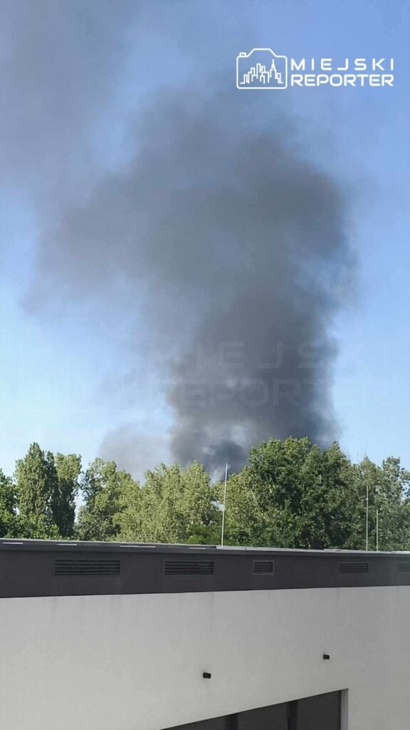 Czarny dym unoszący się nad drzewami w tle, widoczny z poziomu nowoczesnego budynku.
