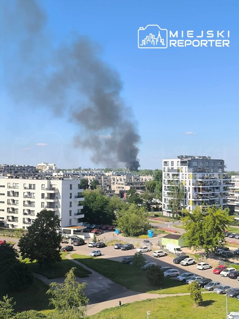 Czarny dym unoszący się nad zabudową mieszkalną w miejskim otoczeniu, w tle widoczne bloki i zieleń.