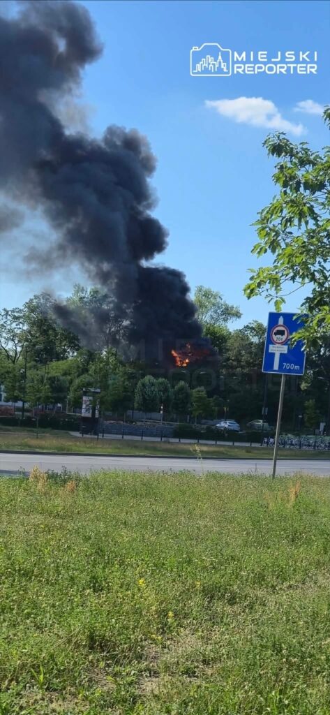 Czarny dym unoszący się z płonącego obiektu w otoczeniu zieleni, przy drodze z oznaczeniem przystanku.