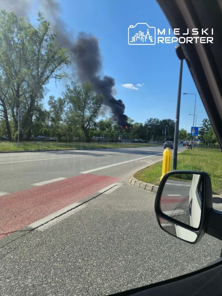 Czarny dym unoszący się z płonącego obiektu w tle, widoczny z drogi, otoczony drzewami i znakami drogowymi.