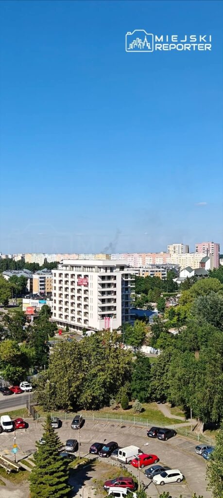 Widok na nowoczesny pięciopiętrowy budynek mieszkalny otoczony zielenią oraz kolorowymi blokami w tle.