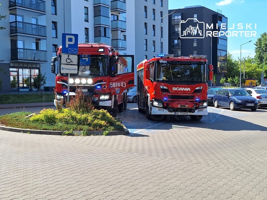Dwa wozy strażackie zaparkowane przed nowoczesnym budynkiem mieszkalnym, otoczone zielenią i miejscami parkingowymi.