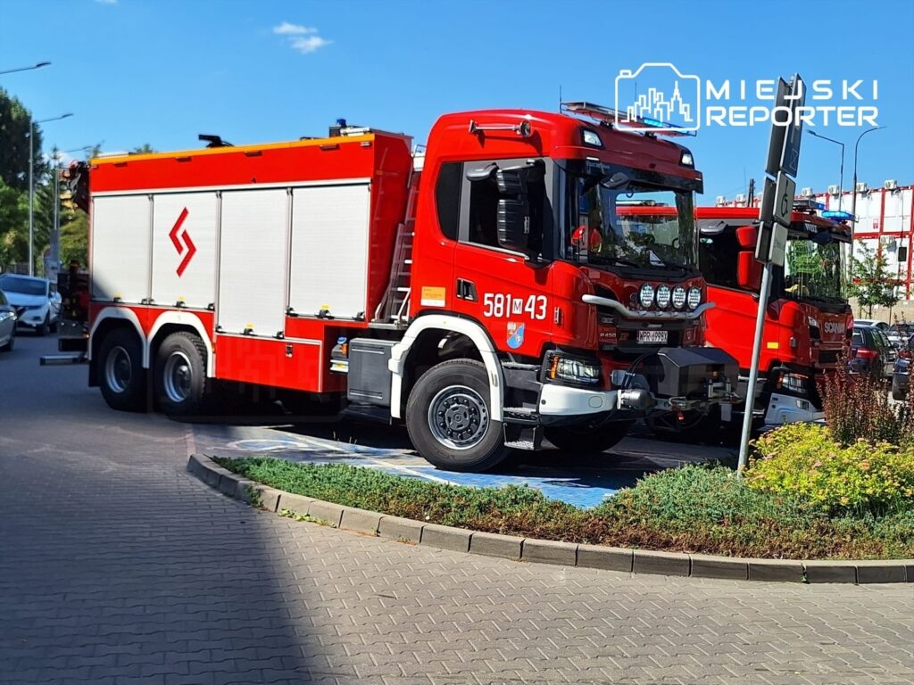 Czerwony wóz strażacki z numerem 581M43 zaparkowany na chodniku, obok drugiego wozu w tle.