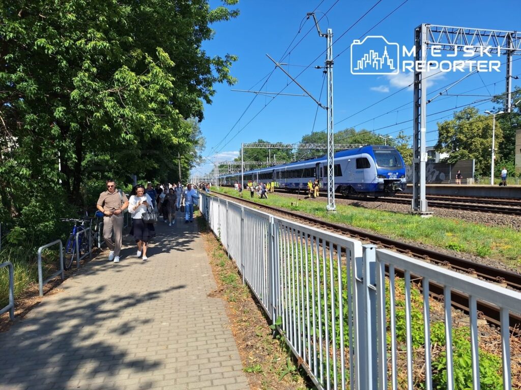 Grupa ludzi spaceruje wzdłuż torów kolejowych, obok nowoczesnego pociągu w niebiesko-srebrnym malowaniu.