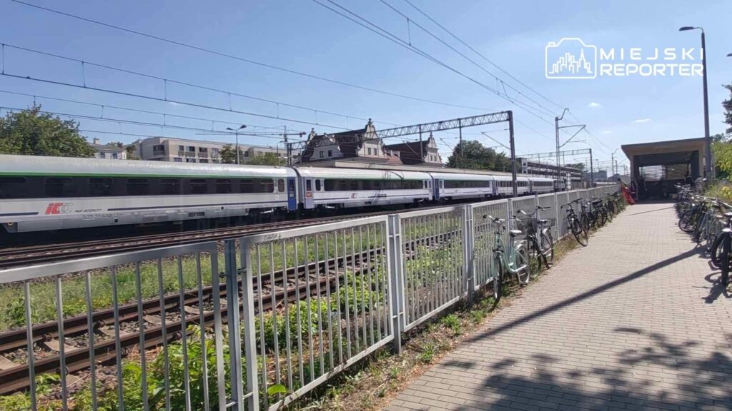 Pociąg pasażerski przejeżdża obok stacji kolejowej, obok której stoją zaparkowane rowery.