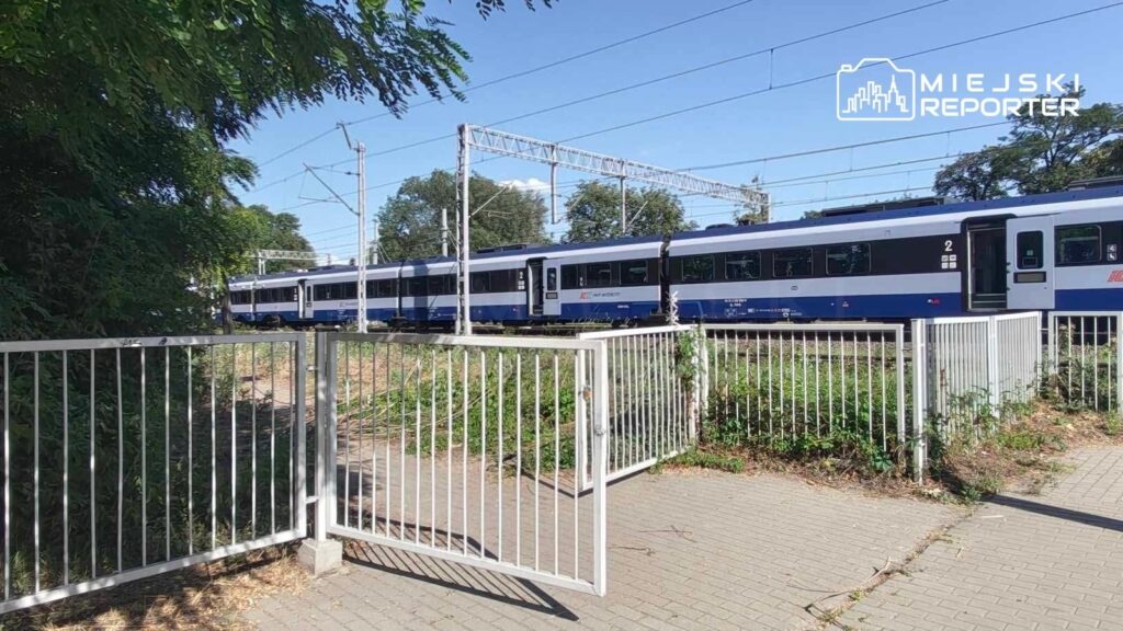 Pociąg osobowy w niebiesko-białych barwach przejeżdża obok otwartych metalowych bramek na tle zieleni.