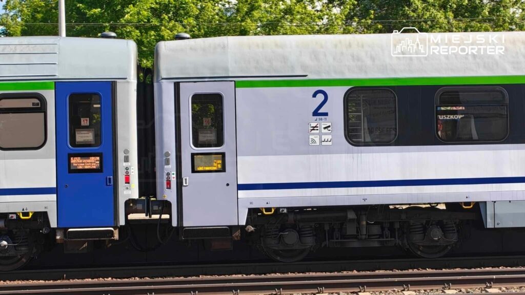 Dwa wagony pociągu z różnymi kolorami i numerami, stojące na torach w otoczeniu zieleni.