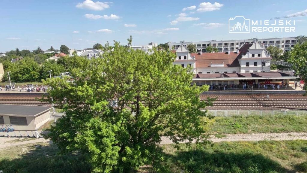 Ludzie czekają na pociąg na peronie, w tle widać dworzec z przeszkloną fasadą i zielone drzewa.
