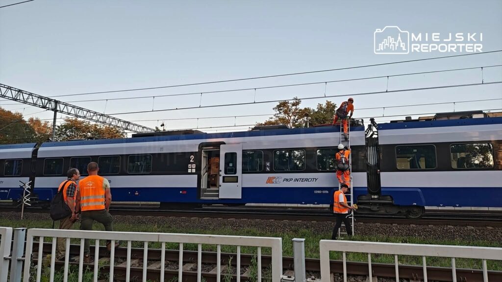 Grupa pracowników w pomarańczowych kamizelkach sprawdza pociąg na torach, jeden z nich wspina się na dach pojazdu.