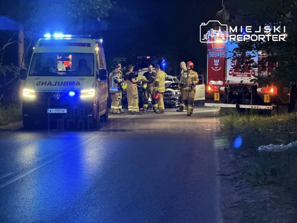 Zespół ratunkowy pracuje przy wypadku drogowym, obok uszkodzonego pojazdu, na oświetlonej ulicy.