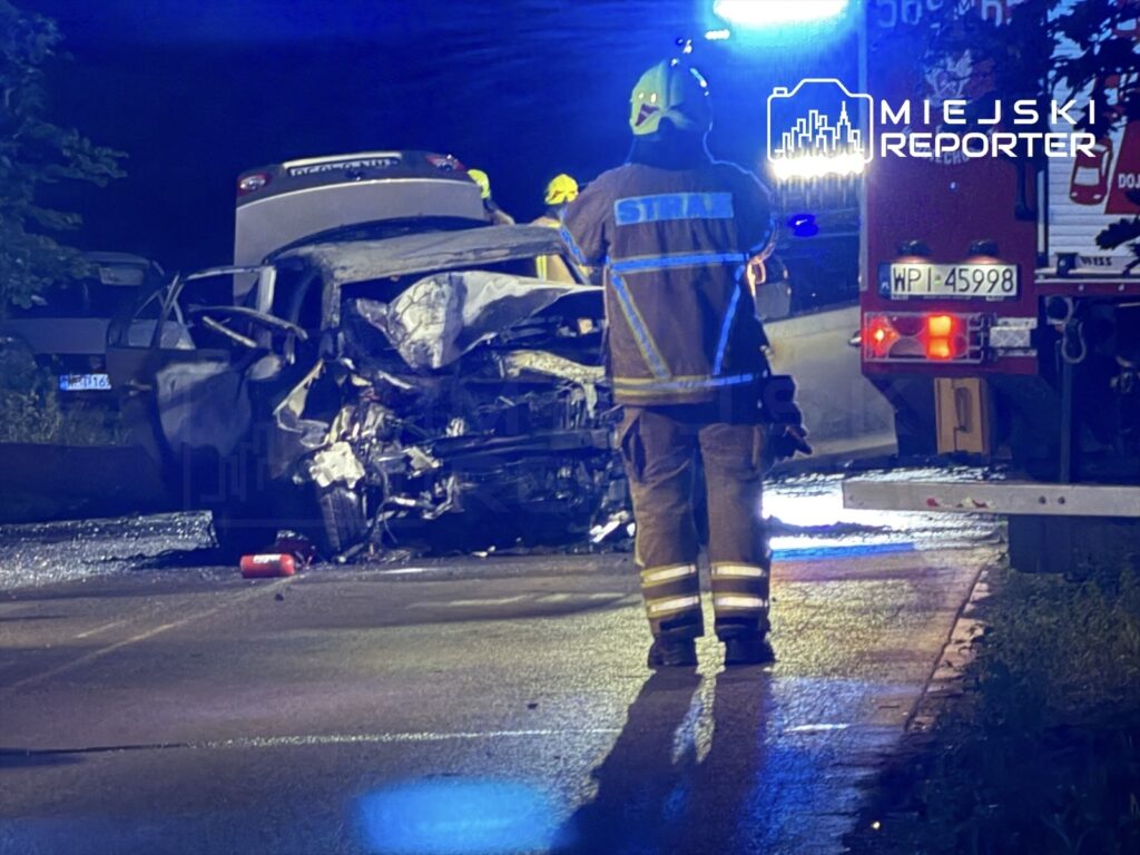 Strażak w hełmie ocenia skutki wypadku drogowego, obok uszkodzonego pojazdu na oświetlonej ulicy.