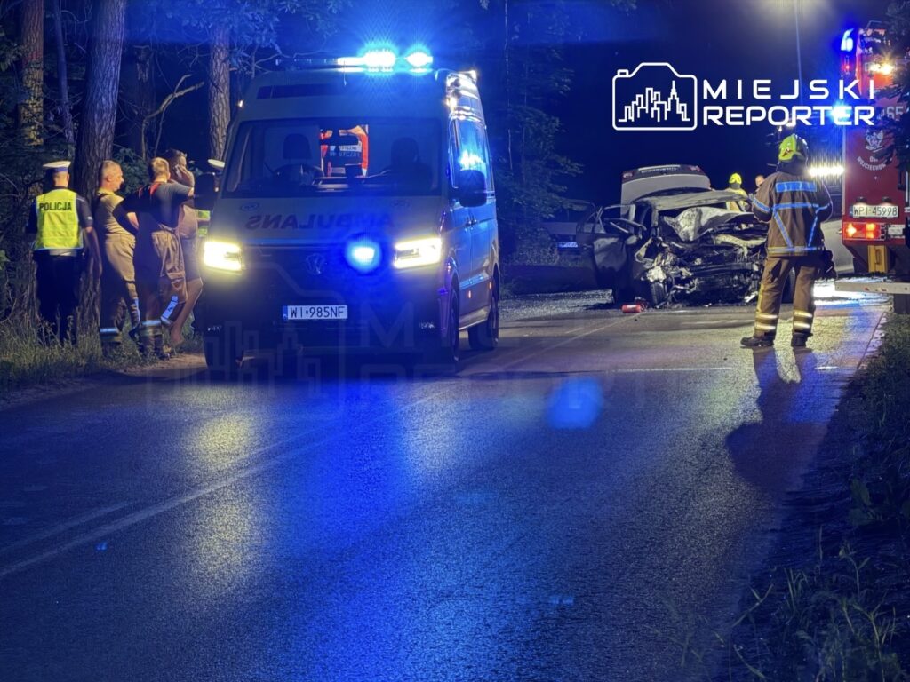 Grupa ratowników i policjantów pracuje przy rozbitym samochodzie na drodze, w tle widać niebieskie światła pojazdów.