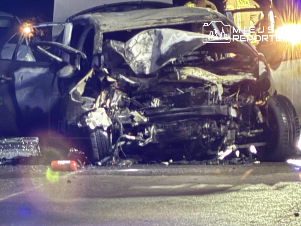 Zniszczony samochód osobowy leży na drodze, obok niego pracują strażacy w mundurach.