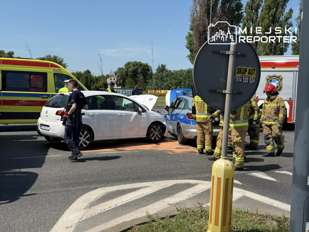 Na skrzyżowaniu dwóch ulic doszło do wypadku samochodowego; policjant i strażacy pomagają uczestnikom.