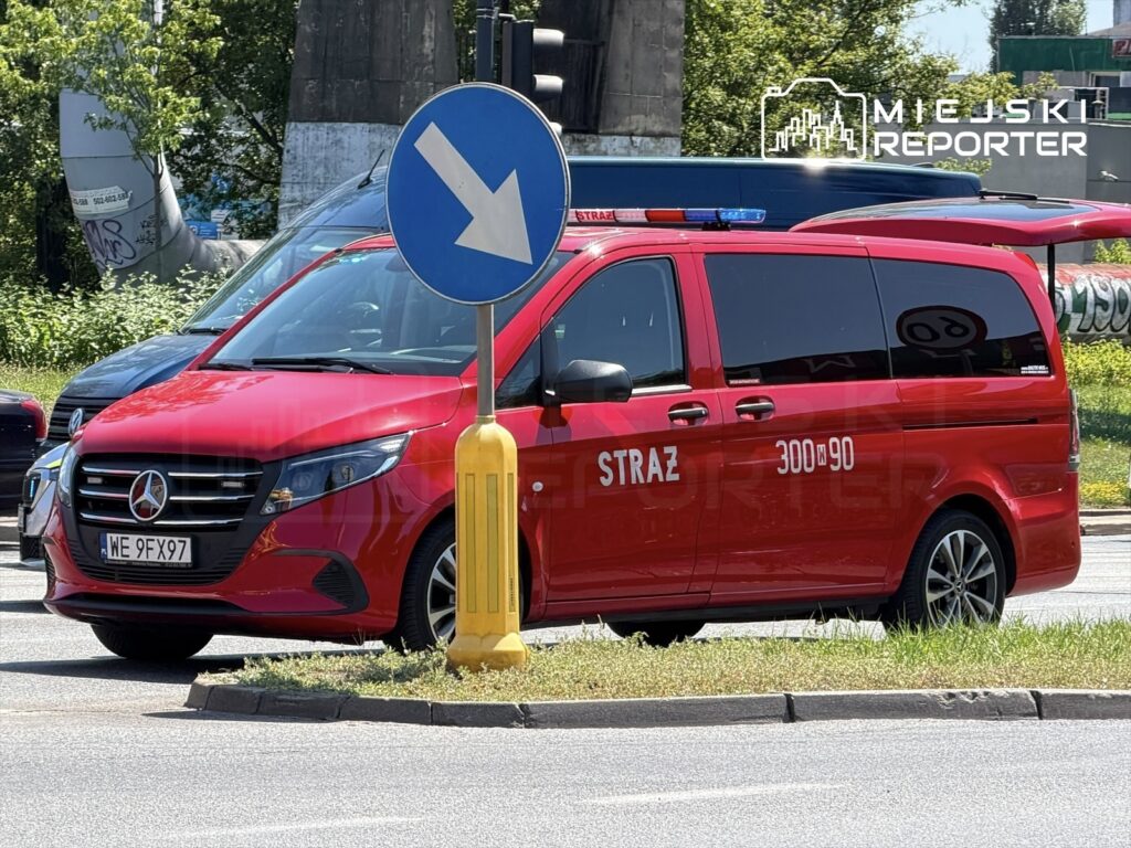 Czerwony samochód straży pożarnej z widocznym napisem "STRAŻ" zatrzymany przy znaku drogowym na skrzyżowaniu.