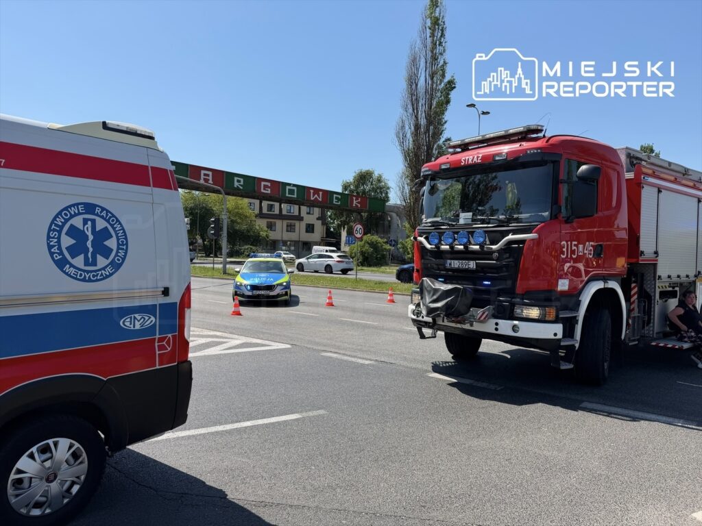 W tle widoczny czerwony wóz strażacki oraz karetka pogotowia, z policją kierującą ruchem na ulicy.