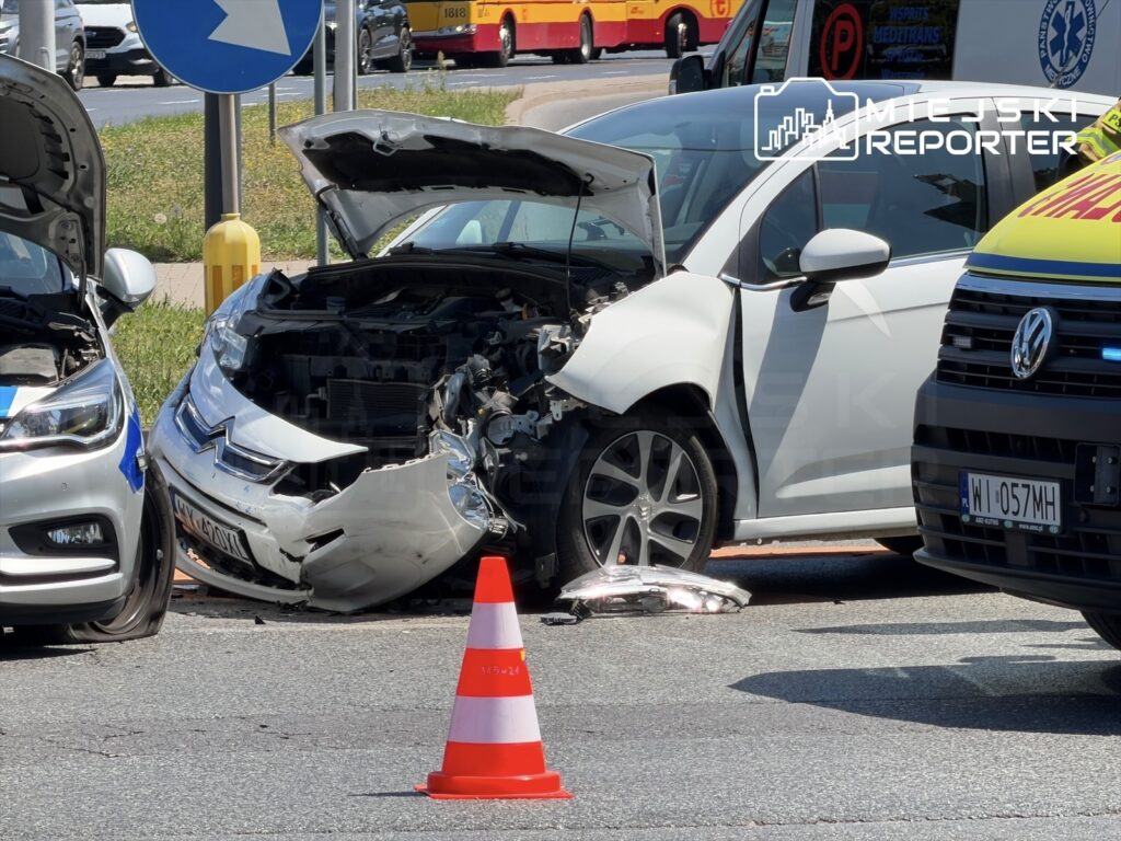 Zderzenie dwóch samochodów osobowych na skrzyżowaniu, z uszkodzoną przednią częścią jednego z pojazdów. W tle widoczne znaki drogowe oraz policyjny radiowóz.