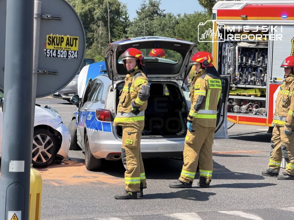 Strażacy w ubraniach ochronnych pracują przy wypadku drogowym, obok radiowozu policji i wozu strażackiego.