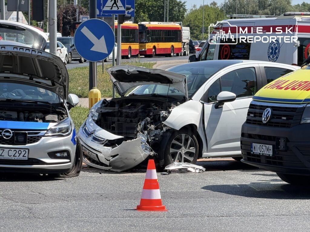Dwa uszkodzone samochody na skrzyżowaniu, z otwartymi maskami, obok czerwony trójkąt ostrzegawczy. W tle widoczne autobusy.