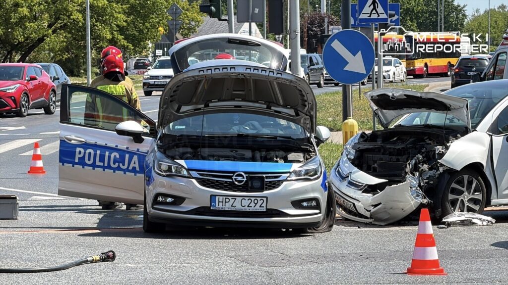 Policjant w żółtej kamizelce ocenia skutki wypadku drogowego na skrzyżowaniu, obok uszkodzonych samochodów.