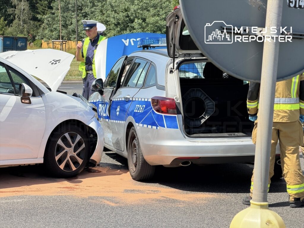 W wyniku kolizji drogowej białe auto osobowe z uszkodzoną maską stoi obok radiowozu policyjnego, a funkcjonariusz rozmawia z kierowcą.