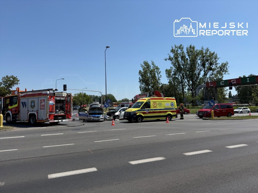 Na skrzyżowaniu stoją pojazdy ratunkowe, w tym straż pożarna i karetka, a policja reguluje ruch po wypadku drogowym.