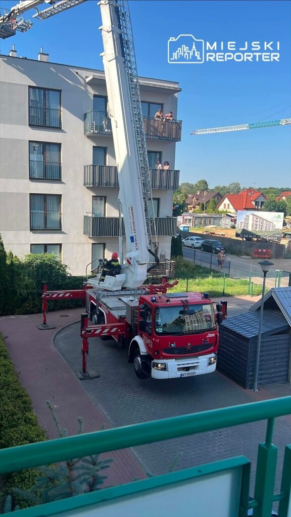Strażak w kabinie wozu strażackiego obsługuje podnośnik, który sięga do balkonów nowoczesnego budynku mieszkalnego.