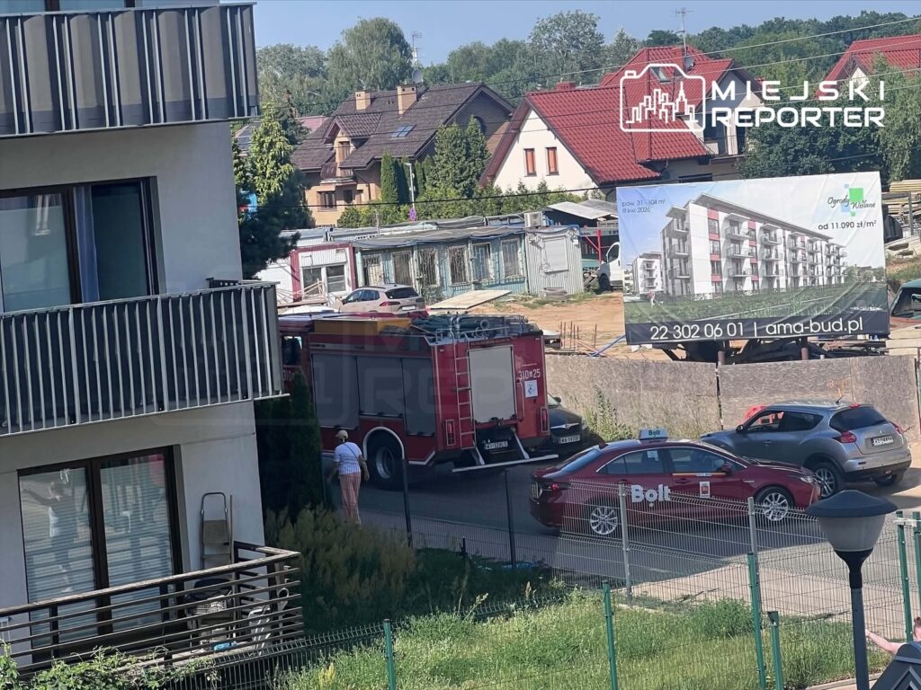 Czerwony wóz strażacki parkuje obok budowy, a obok niego stoi mężczyzna w białej koszuli i kobieta w różowych spodniach.