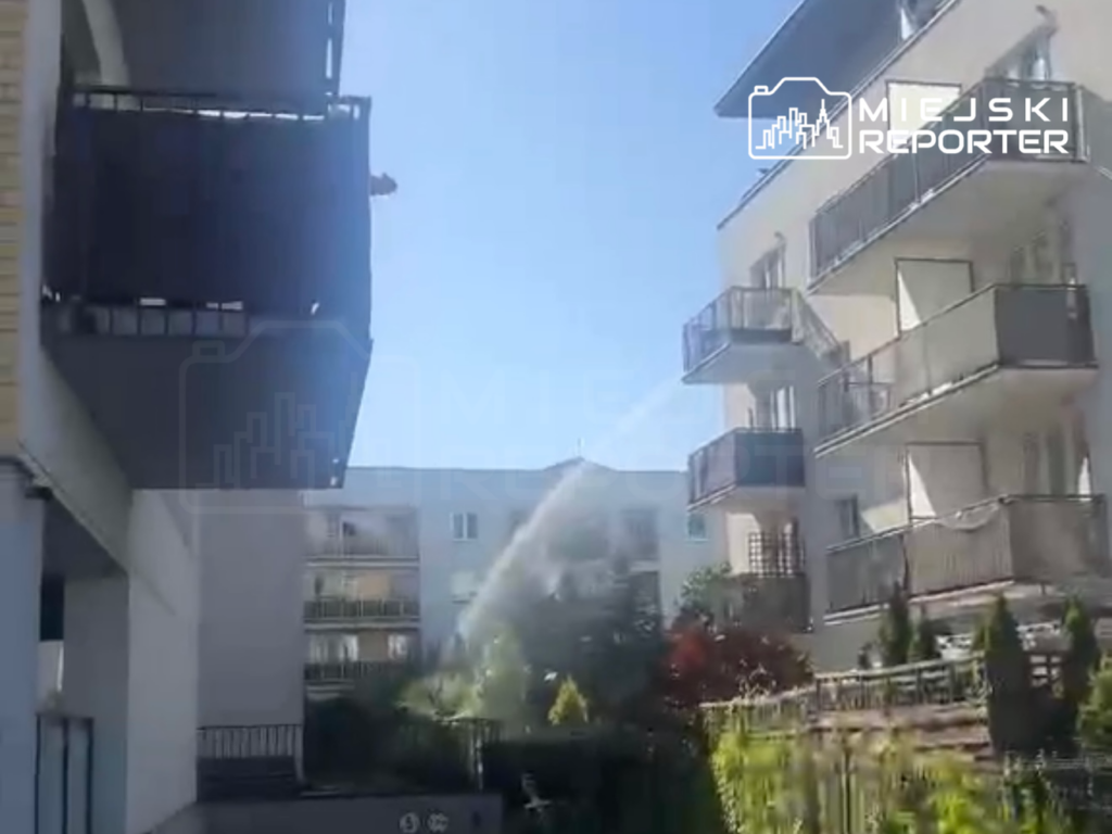 Mężczyzna na balkonie obserwujący strumień wody w ogrodzie między nowoczesnymi budynkami mieszkalnymi.