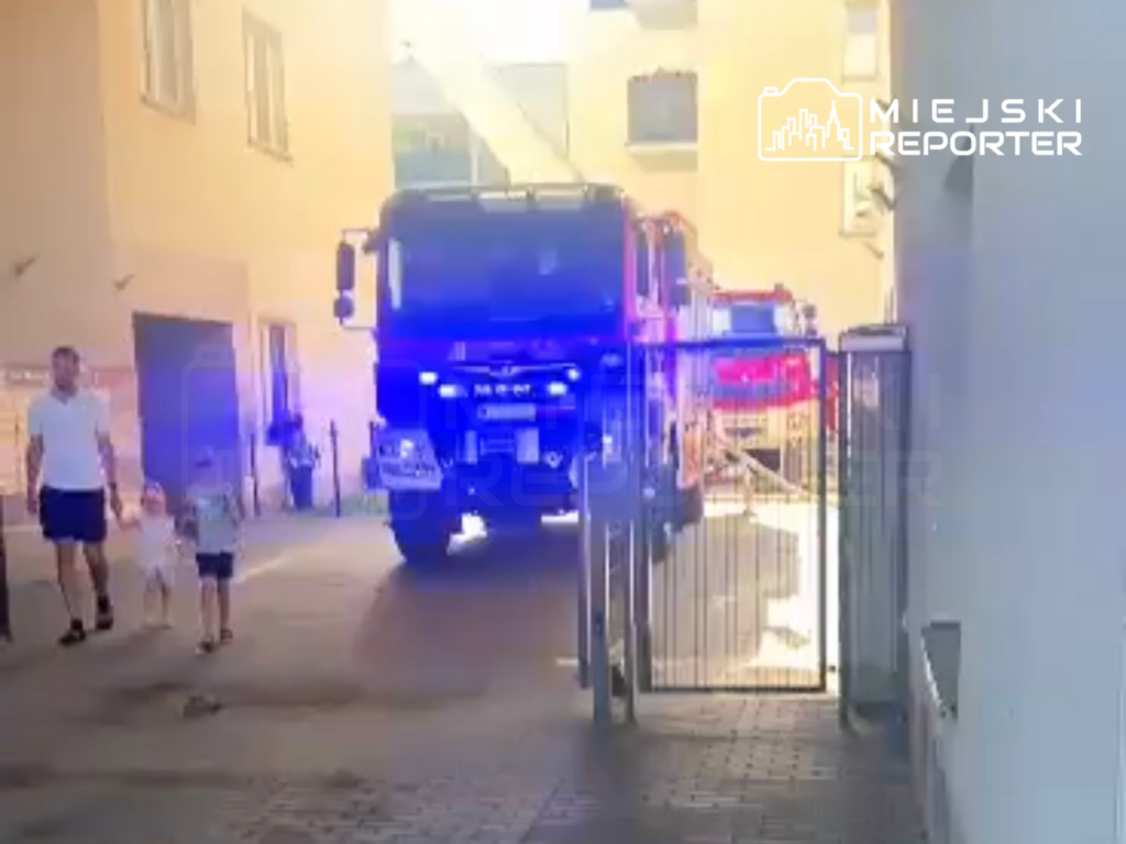 Mężczyzna trzymający dwoje dzieci spaceruje obok wozu strażackiego z włączonymi sygnalami świetlnymi w wąskim podwórku.