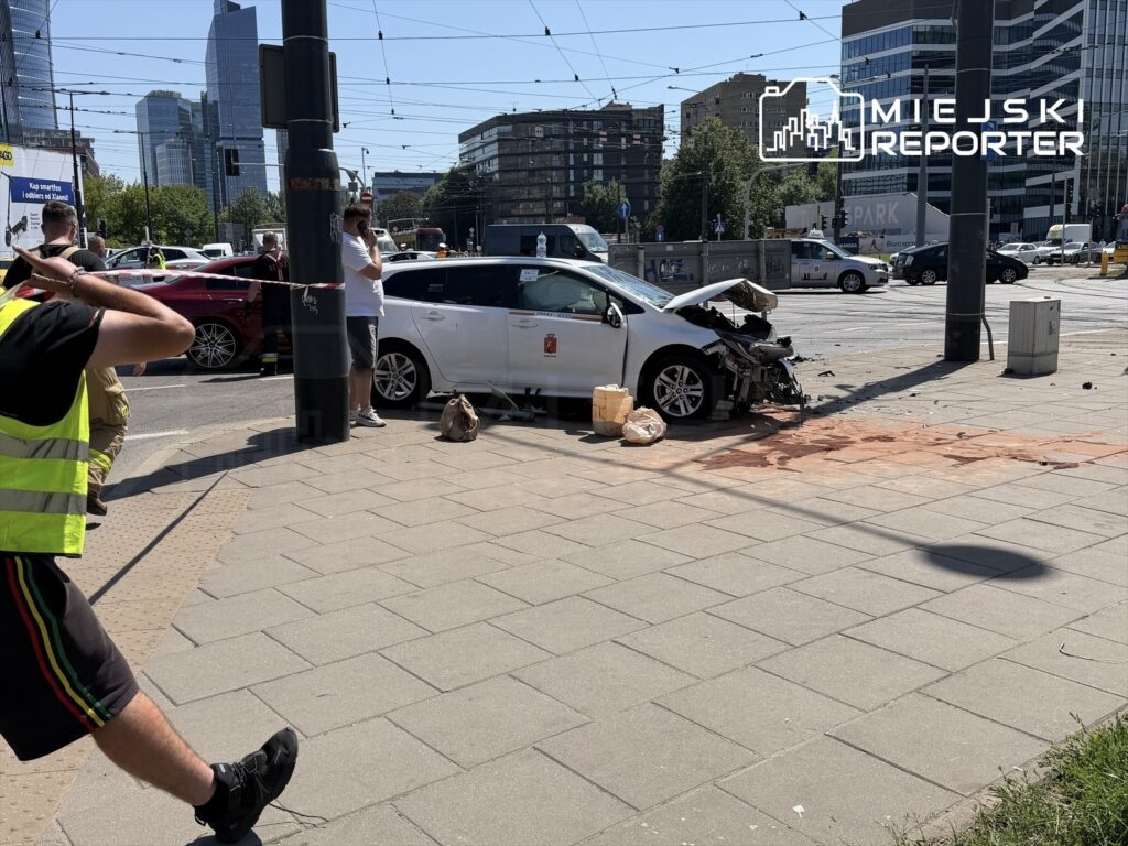 Wypadek drogowy z uszkodzonym samochodem osobowym na skrzyżowaniu, obok grupy osób w odblaskowych kamizelkach.