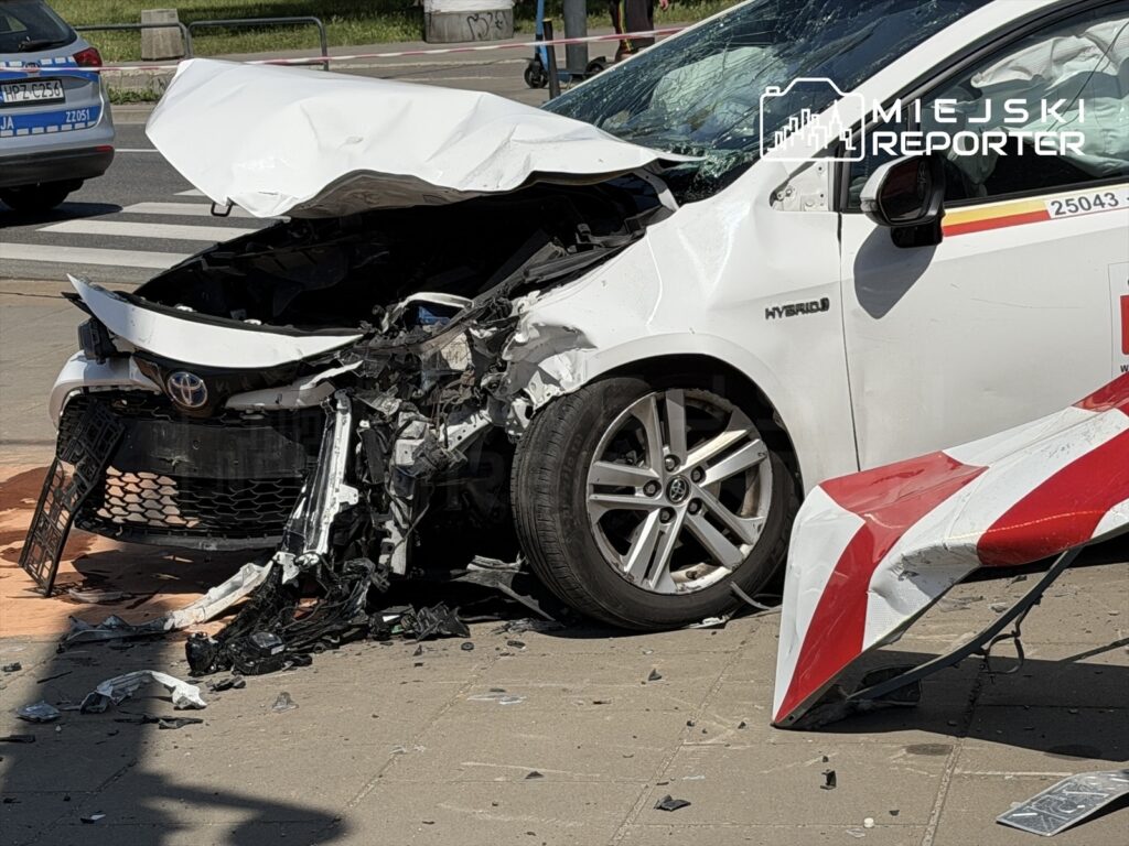 Zniszczony biały samochód osobowy z uszkodzoną przednią częścią stoi na chodniku, wokół rozrzucone fragmenty pojazdu.