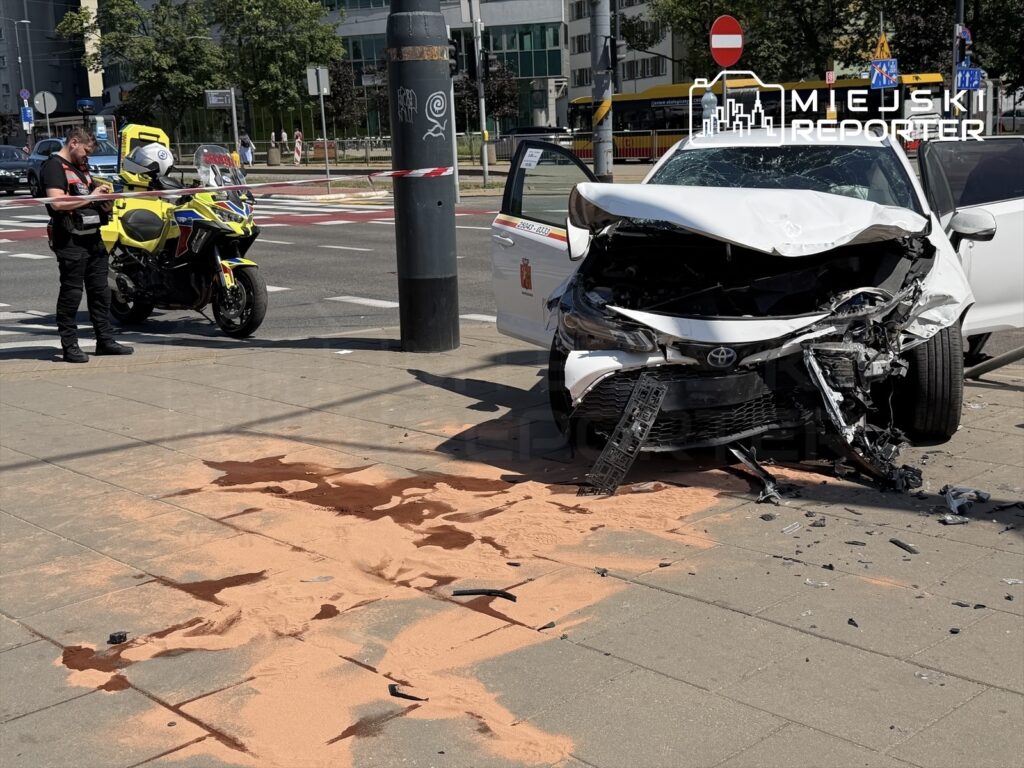 W miejscu wypadku leży uszkodzony samochód osobowy z rozbitą przednią częścią, obok policjant z aparatem fotograficznym. Na jezdni widoczny jest rozlany ciecz i piasek.