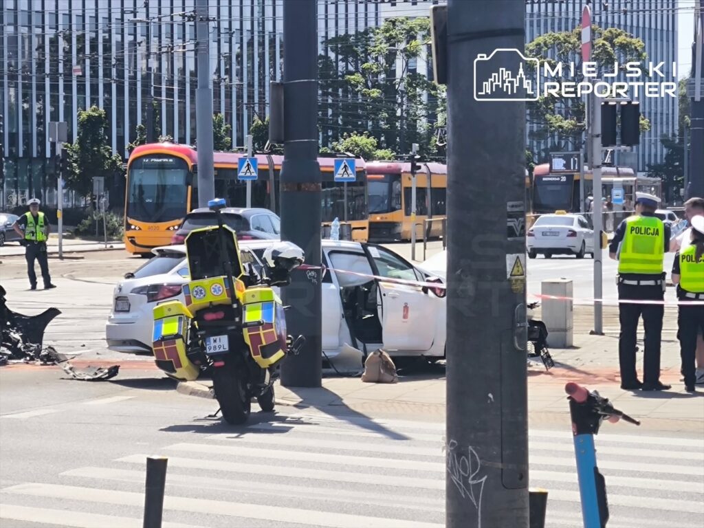 Na skrzyżowaniu stoją policjanci, obok uszkodzone samochody oraz motocykl służby. W tle widać tramwaj.