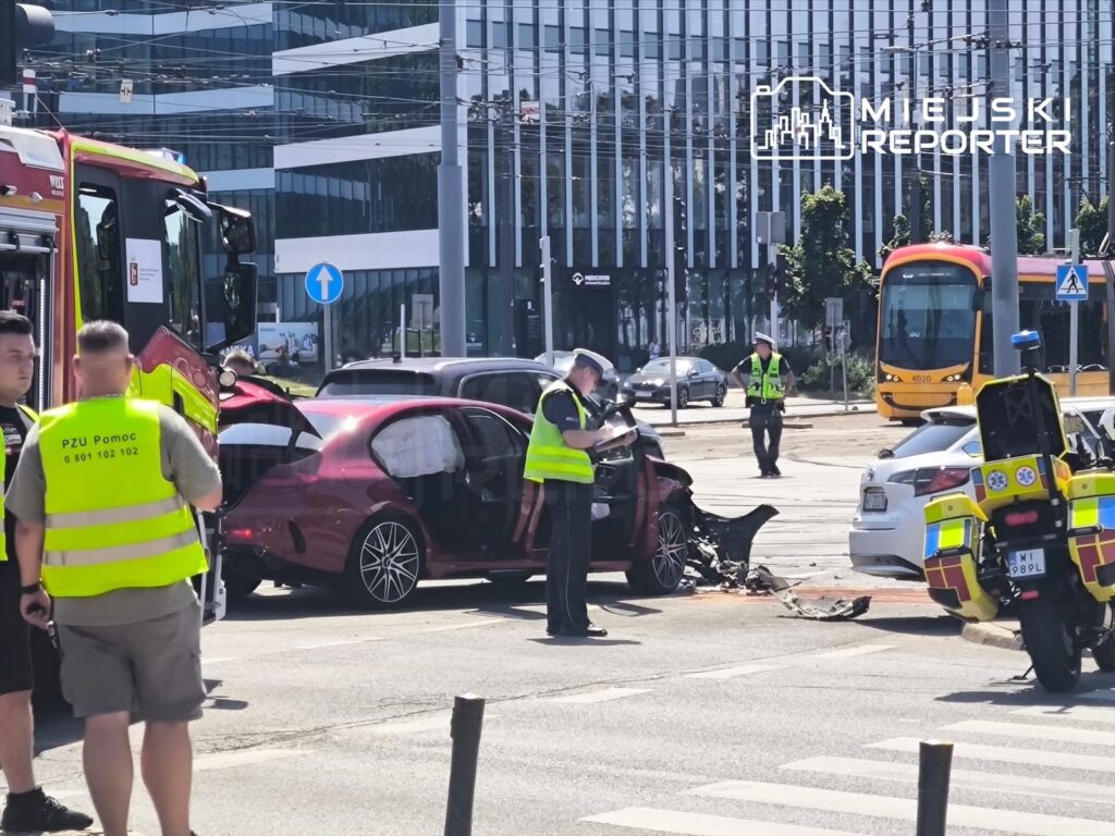 Na skrzyżowaniu, strażacy i policjanci badają miejsce wypadku drogowego z udziałem uszkodzonego czerwonego samochodu.