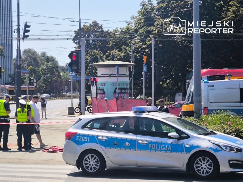 Policjanci w kamizelkach odblaskowych zabezpieczają miejsce zdarzenia przy czerwonym świetle, obok radiowozu.