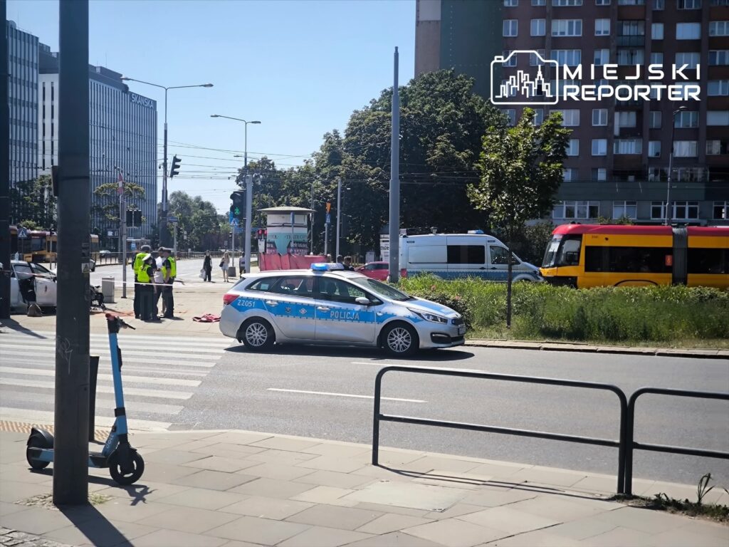 Policjanci w kamizelkach odblaskowych prowadzą czynności na skrzyżowaniu, obok radiowozu policyjnego.