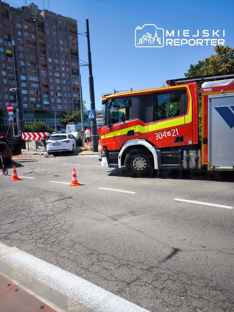 Czerwony wóz strażacki z numerem 304W21 stoi na drodze, w tle widoczny biały samochód oraz blok mieszkalny.