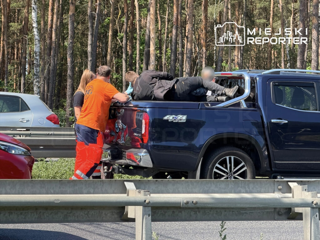 Mężczyzna w pomarańczowej koszulce oraz kobieta pomagają osobie leżącej w skrzyni niebieskiego pickupa na drodze.