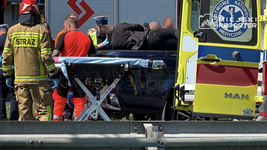 Ratownicy medyczni udzielają pomocy mężczyźnie leżącemu na samochodzie, obok stoi strażak w mundurze.