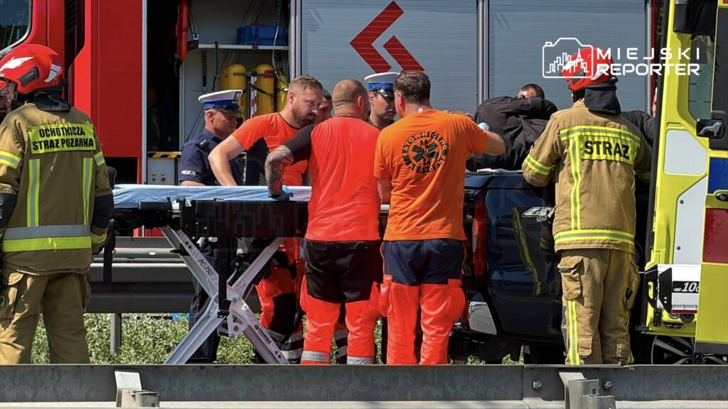 Grupa ratowników w pomarańczowych i żółtych mundurach transportuje poszkodowanego na noszach obok pojazdów służb ratunkowych.