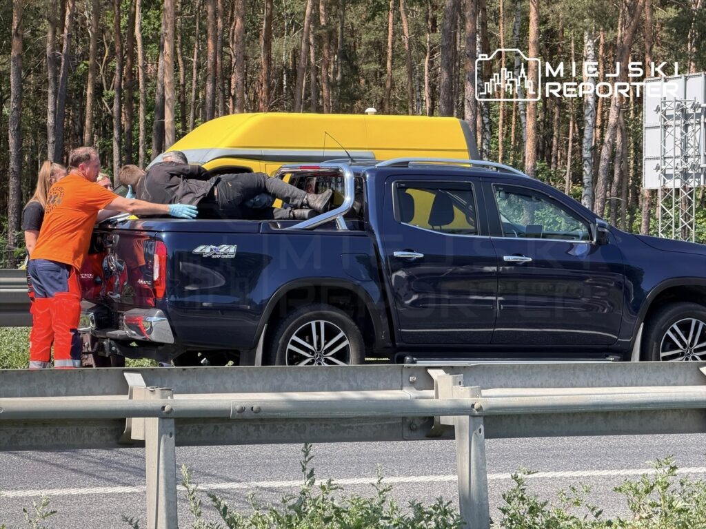 Grupa ratowników udziela pomocy mężczyźnie leżącemu w tylnej części pickup'a na drodze, w otoczeniu lasu.
