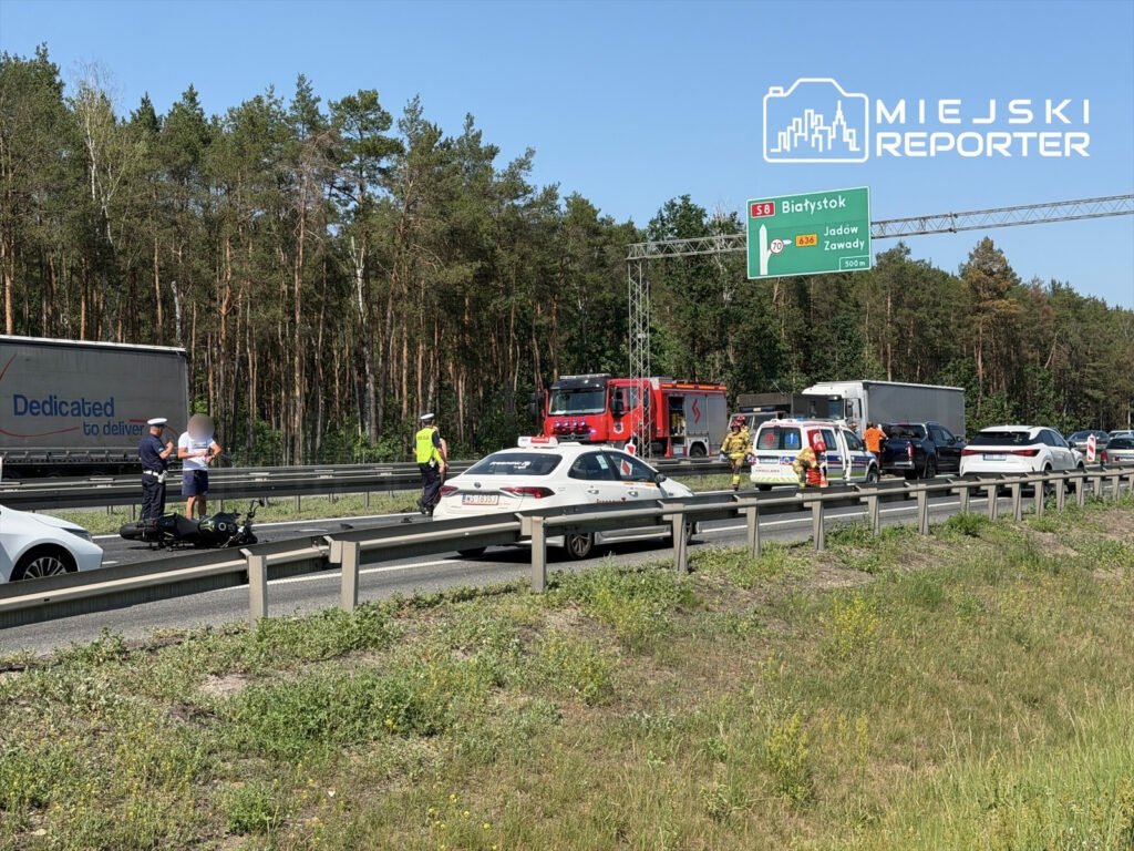 Na drodze stoją policjanci rozmawiający z motocyklistą obok przewróconego motocykla, w tle widać pojazdy ratunkowe.