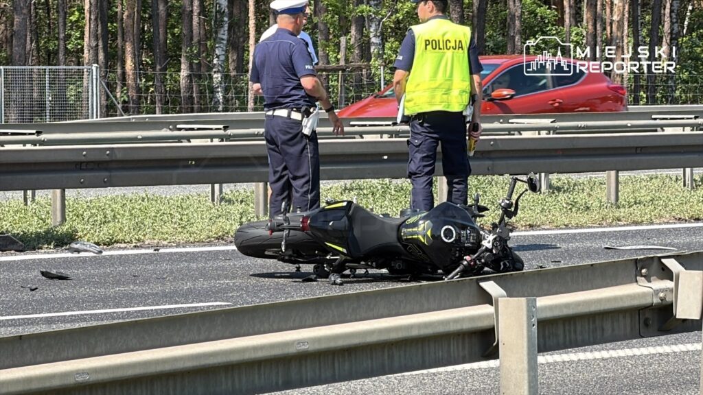 Trzech policjantów bada przewrócony motocykl na pasie drogi, w tle widać zieleń drzew i przejeżdżający samochód.