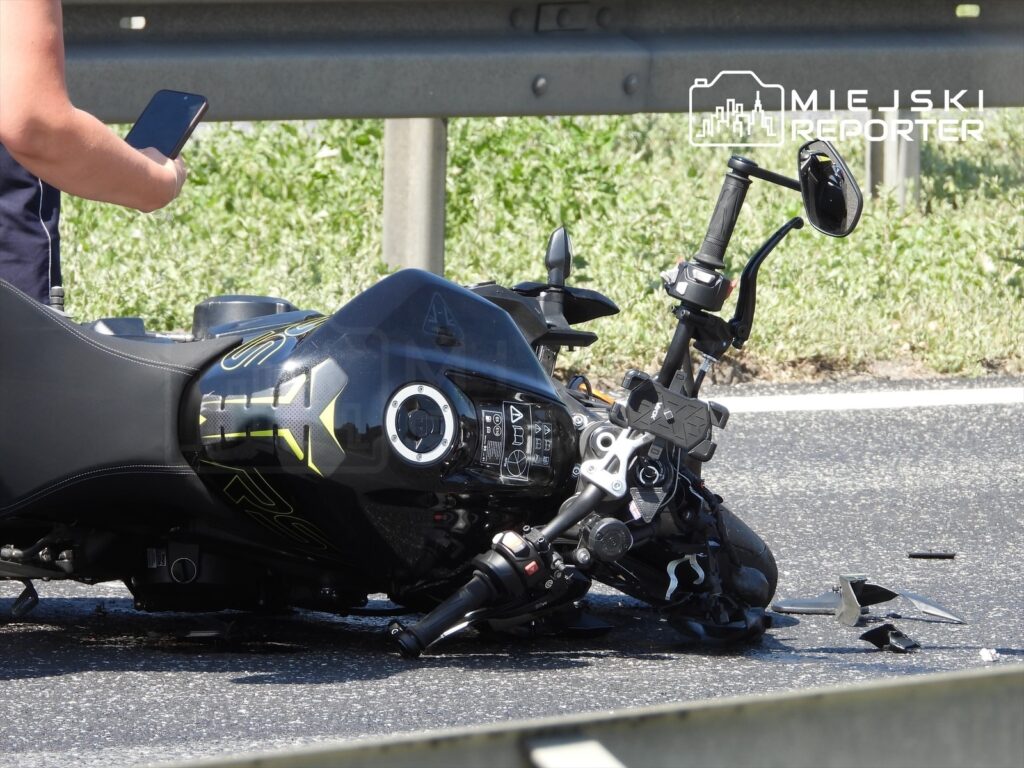 Mężczyzna trzymający telefon stoi obok przewróconego motocykla na drodze, otoczonego odłamkami.