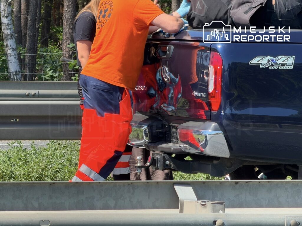 Mężczyzna w pomarańczowej koszulce pomaga przy naprawie niebieskiego samochodu dostawczego na poboczu drogi.