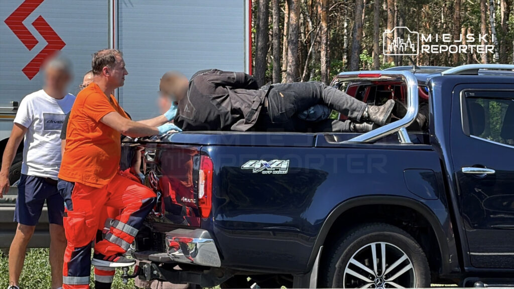 Ratownik medyczny w pomarańczowym stroju udziela pomocy osobie leżącej w tylnej części pickup'a w lesie.